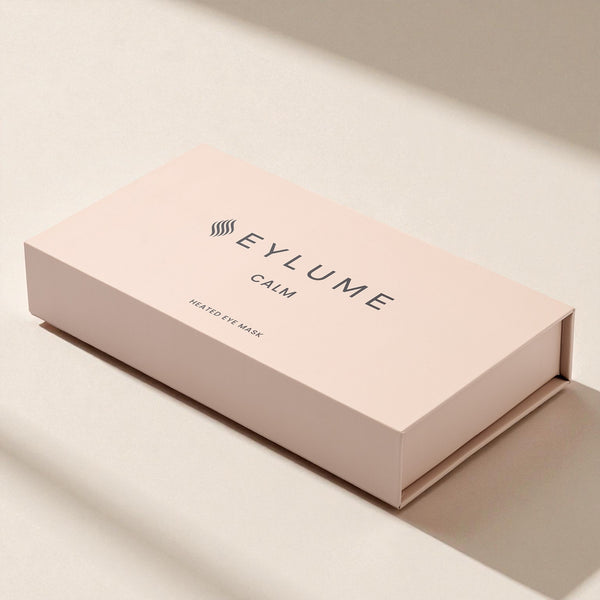 EYLUME Calm Heated Eye Mask box on a warm beige surface under soft natural sunlight, creating gentle shadows and a calm, premium aesthetic.